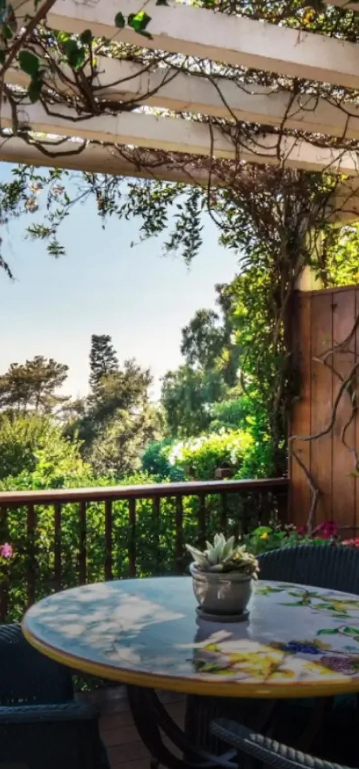 A private outdoor patio at San Ysidro Ranch featuring a decorative tiled table under a white pergola covered in climbing vines. Lush green trees and a clear blue sky are visible beyond the wooden railing. - In Beverly Hills, CA