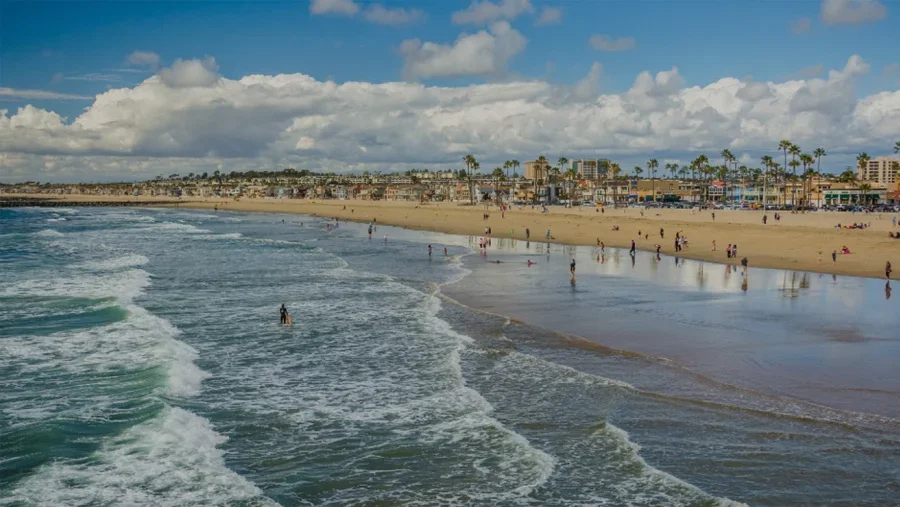 A wide-angle view of a crowded sandy beach with gentle waves crashing on the shore. Palm trees and coastal buildings line the background under a blue sky with fluffy white clouds. - location in Beverly Hills, CA