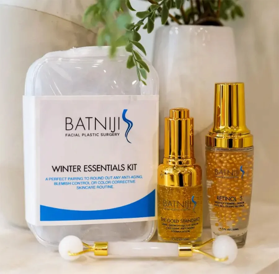 A collection of Batniji Facial Plastic Surgery skincare products, including a "Winter Essentials Kit" and two gold-capped serum bottles labeled "The Gold Standard" and "Retinol .5," accompanied by a white jade facial roller. - Skincare in Beverly Hills, CA