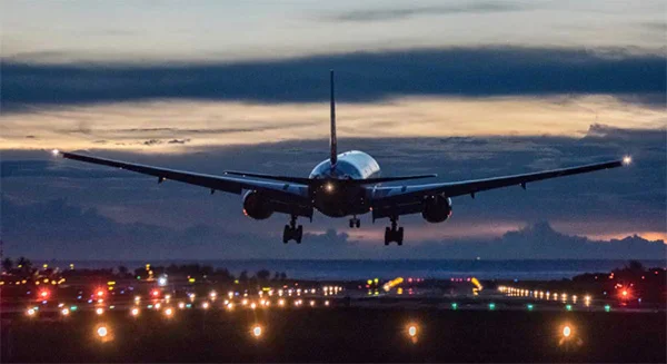 A rear view of a large passenger jet descending toward an airport runway at twilight. The landing gear is down, and the plane's silhouette is set against a dark, moody sky with a faint orange glow on the horizon. Below, the runway is illuminated by rows of glowing white and orange navigation lights. - Travel airports in Beverly Hills, CA