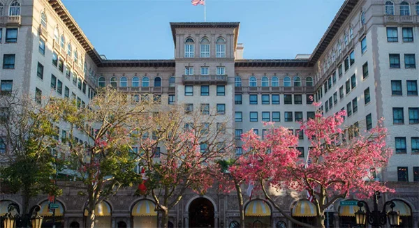 A wide-angle shot of a grand, multi-story historic hotel building with classic European-style architecture. In the foreground, vibrant pink cherry blossoms and green trees line the street. Yellow-and-white striped awnings cover the ground-floor windows, and an American flag flies from the central roof tower. - Travel in Beverly Hills, CA
