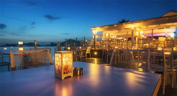 A peaceful evening scene at an outdoor waterfront restaurant. In the foreground, a modern white lantern with a circular cutout pattern glows warmly on a table. In the background, white patio chairs and tables are arranged under a brightly lit canopy, all overlooking a calm ocean under a deep blue dusk sky. - Traavel in Beverly Hills, CA