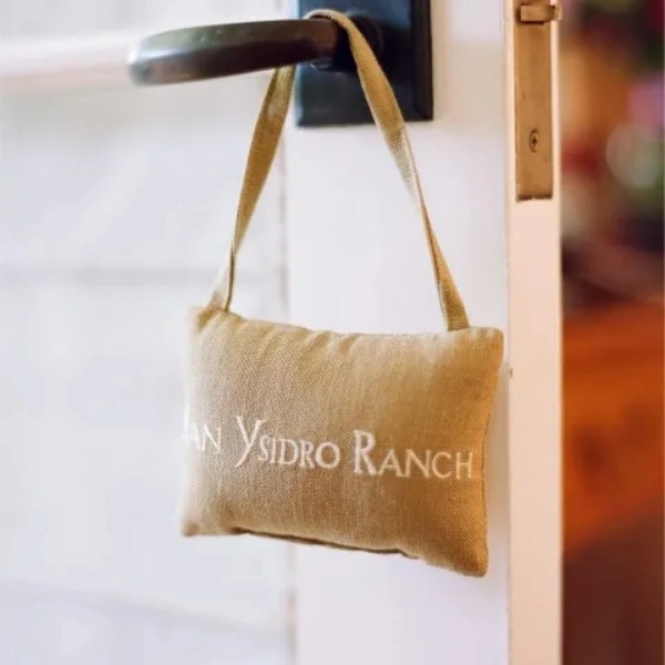 A small, tan burlap pillow with the words "San Ysidro Ranch" embroidered in white hangs from a dark metal door handle. The background shows a white door frame and a blurred interior, suggesting a luxury hospitality or recovery suite setting. - In Beverly Hills, CA
