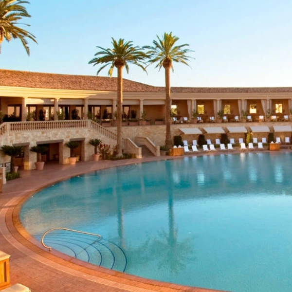 A wide shot of a luxury resort pool area under a clear blue sky. The large, curved turquoise pool is surrounded by a terracotta patio with white lounge chairs and tall palm trees. In the background, a Mediterranean-style building features elegant stone arches and a tiled roof. - In Beverly Hills, CA