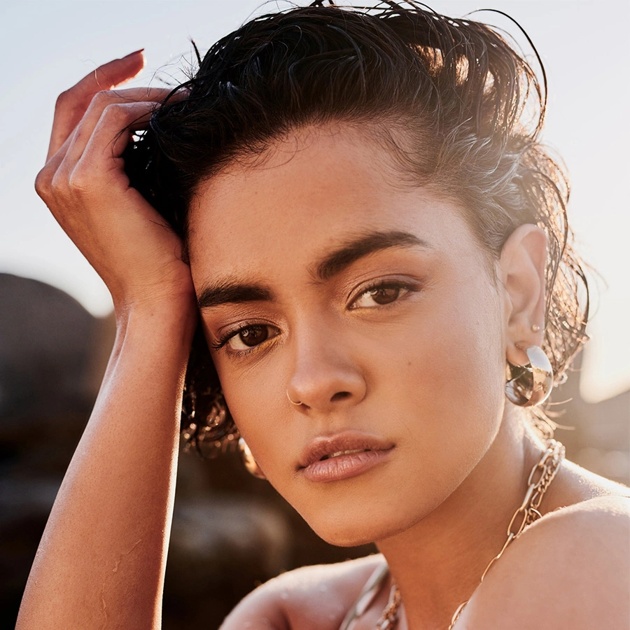 A close-up, front-facing portrait of a woman with short, dark, textured hair. She is wearing large silver hoop earrings and a silver chain necklace. The warm sun is behind her, creating a soft glow around her silhouette. - Brow lift in Beverly Hills, CA