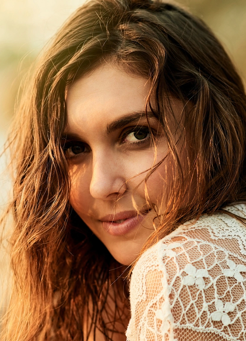 A close-up, intimate portrait of a woman with wavy, sun-kissed hair and a gentle smile. She is wearing a white lace garment. The warm evening light catches the strands of her hair and highlights the smooth, elevated area of her brow and forehead. - Brow lift in Beverly Hills, CA