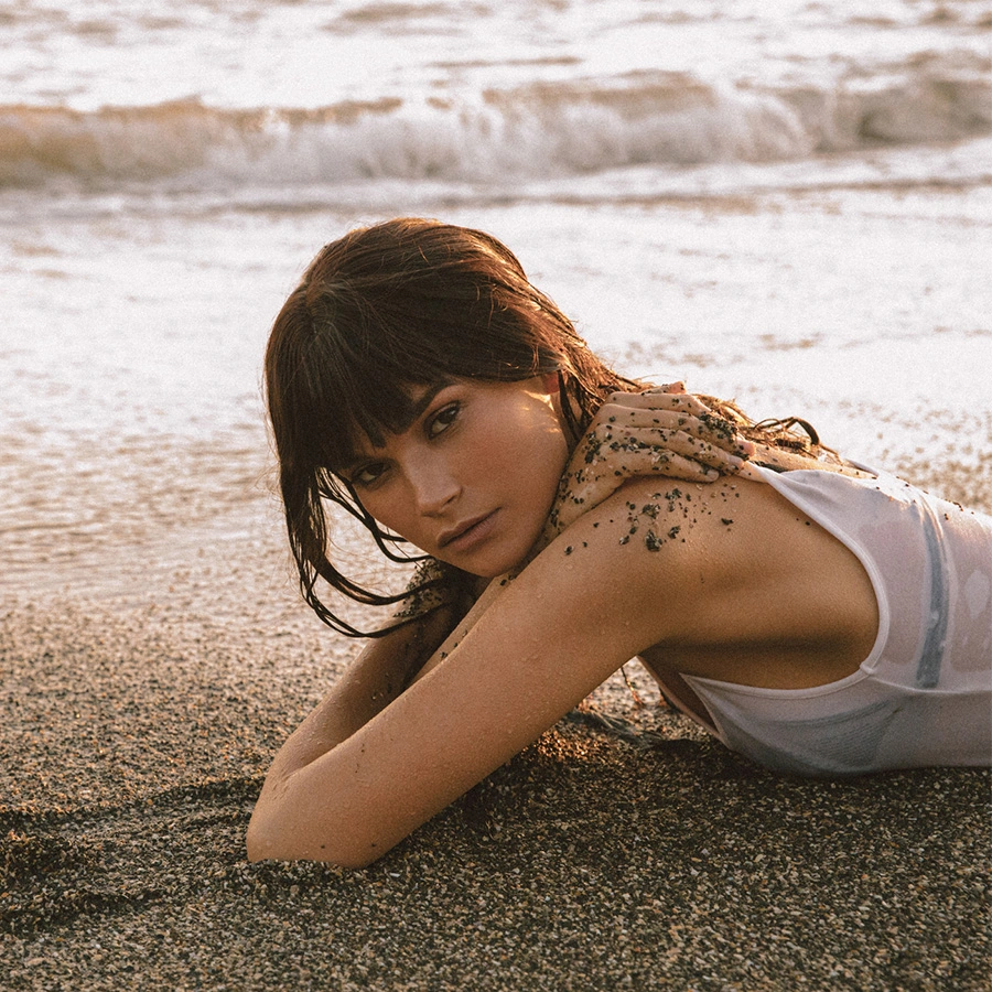A woman with dark hair and bangs lying on a sandy beach as waves crash behind her. She is wearing a white tank top, and her shoulder is lightly dusted with sand. She is looking intensely toward the camera. - Buccal Fat Reduction in Beverly Hills, CA
