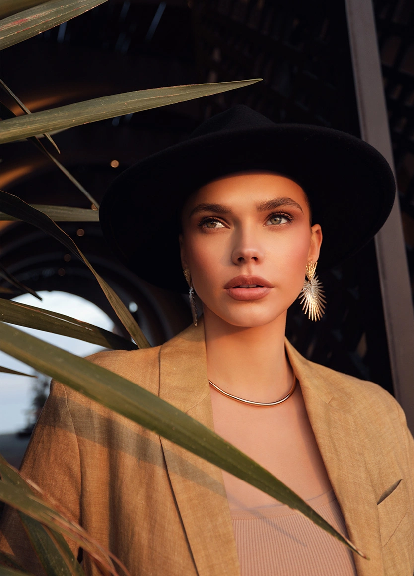 A sophisticated portrait of a woman wearing a tan blazer, a black wide-brimmed hat, and large starburst-shaped earrings. She gazes upward and to the side with a neutral expression. Warm, low-angled sunlight casts shadows from nearby palm leaves across her face, highlighting her chiseled cheekbones and defined facial structure. - Buccal fat reduction in Beverly Hills, CA