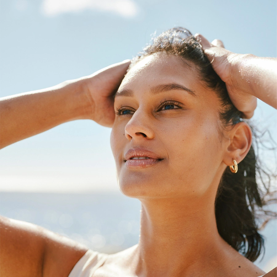 A woman with her hair pulled back in a ponytail, looking upward with a clear, bright expression. Her hands are resting on the back of her head, and she is wearing small gold hoop earrings. The background shows a bright, blurry seascape. - Eyelid surgery in Beverly Hills, CA