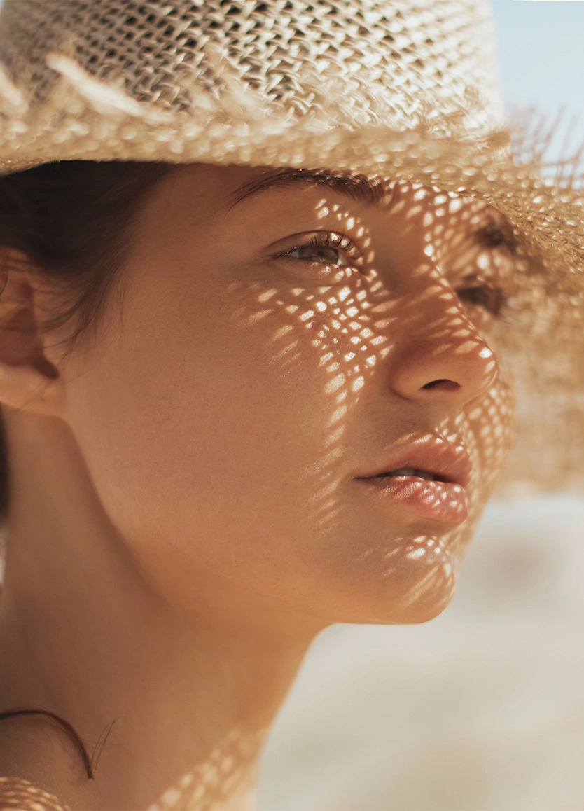 A close-up profile of a woman’s face, focusing on her eyes and cheek. She is wearing a wide-brimmed straw hat that casts a tight, mesh-like shadow pattern across her face. The warm sunlight emphasizes her clear complexion and the delicate area around her eyes. - Eyelid surgery in Beverly Hills, CA