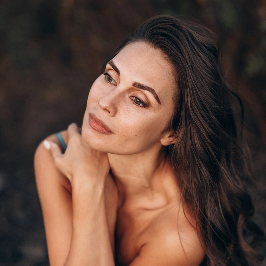 A portrait of a woman with long, dark wavy hair looking upward and to the side with a serene expression. She has her hands crossed near her neck and shoulders. The background is dark and out of focus. - Facelift in Beverly Hills, CA