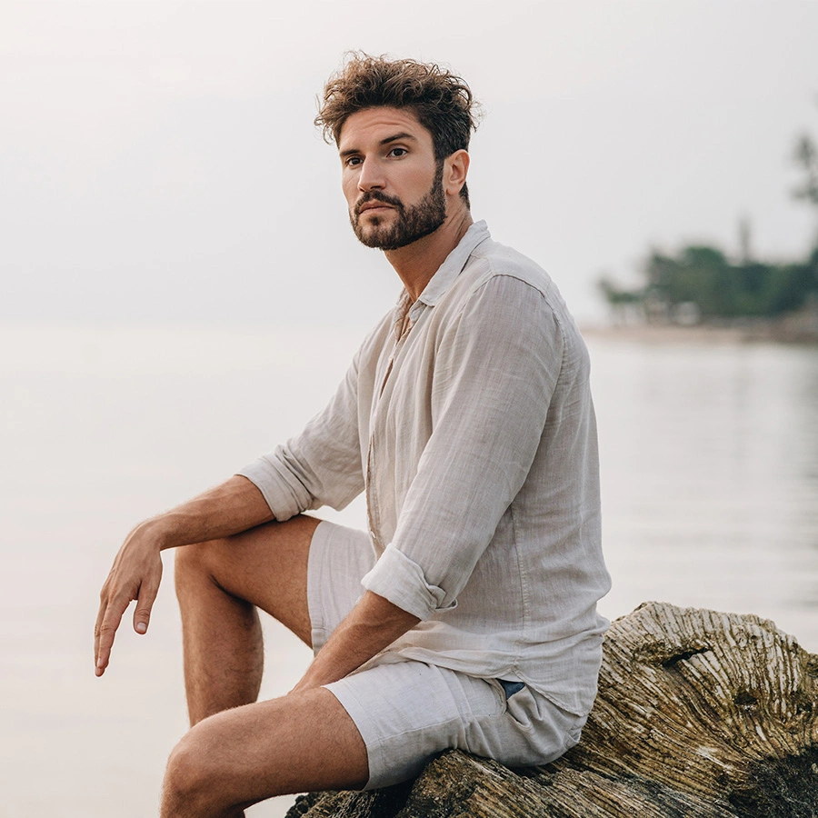 A bearded man with curly hair wears a light linen shirt and shorts, sitting on a large piece of driftwood by a calm, misty shore. - Male facial plastic surgery in Beverly Hills, CA