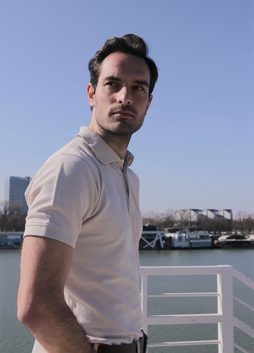 A man in a beige polo shirt standing on a boat or pier, looking off into the distance. He has a strong jawline and short dark hair, with a river and city skyline in the clear, blue-sky background. - Male facial plastic surgery in Beverly Hills, CA