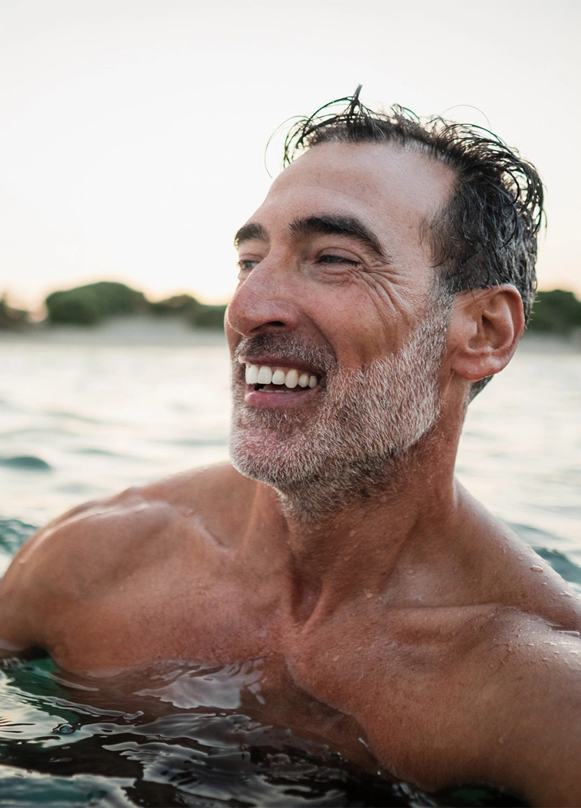 A smiling, middle-aged man with salt-and-pepper hair and a beard, partially submerged in the ocean. The photo is taken at dusk, showing a healthy, happy expression and clear skin on his face and chest. - Mohs Facial Reconstruction in Beverly Hills, CA