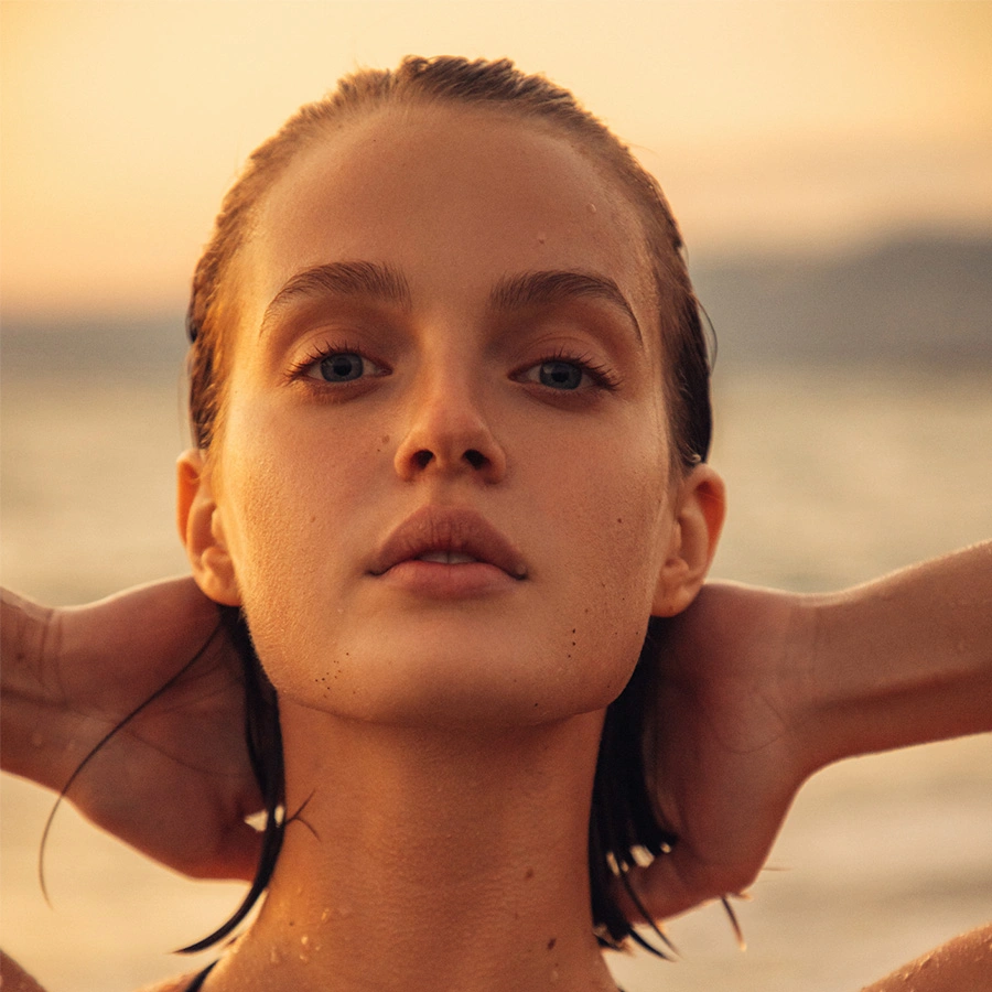 A close-up, front-facing portrait of a woman with slicked-back wet hair and blue eyes, holding her hands behind her neck against a sunset ocean backdrop. - Neck Liposuction in Beverly Hills, CA