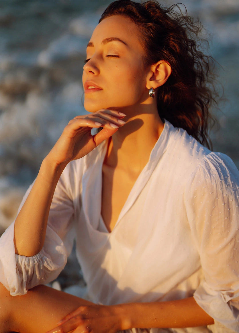 A woman with her eyes closed and chin tilted up, soaking in the golden hour light. She is wearing a white, semi-sheer blouse and small silver hoop earrings. The warm sunset light creates a sharp, clean silhouette of her neck and jawline. - Neck Liposuction in Beverly Hills, CA