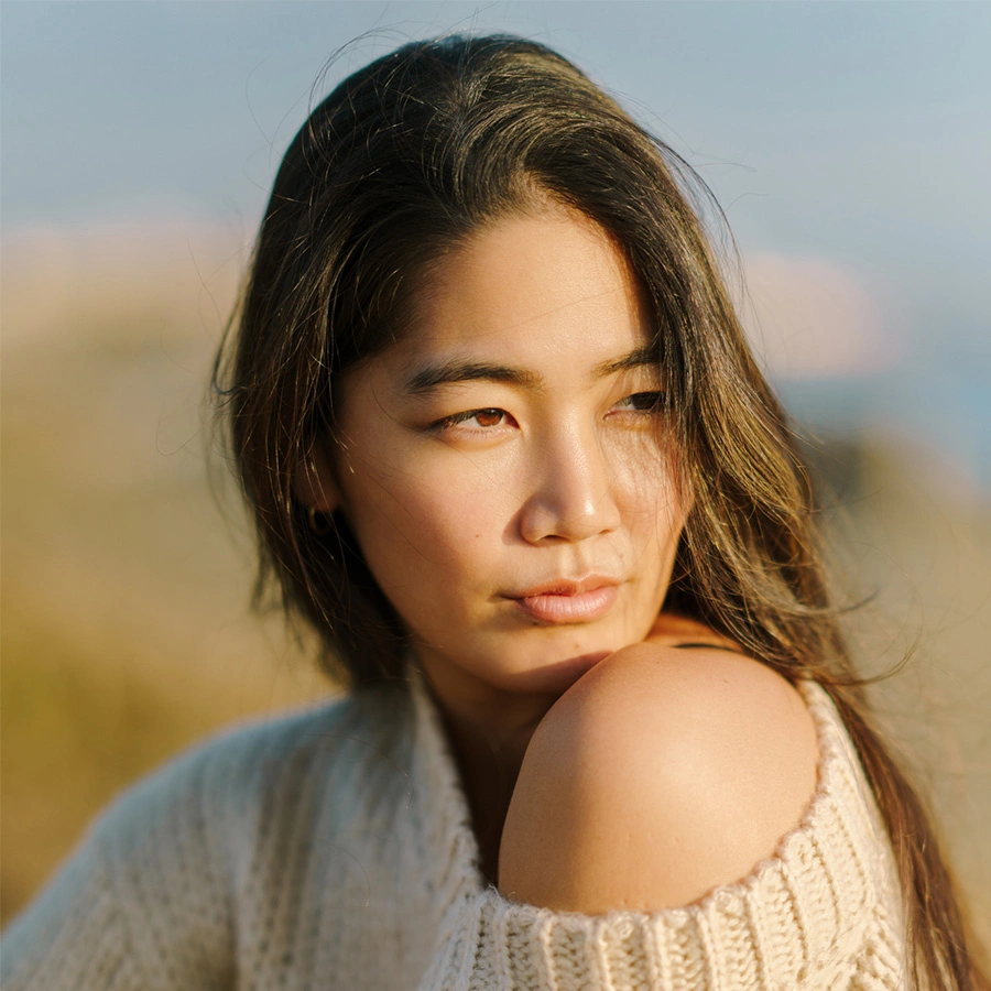 A woman with long dark hair looks off to the side, wearing a cream-colored off-the-shoulder knit sweater in a soft, natural outdoor setting. - Non surgical Facelift in Beverly Hills, CA