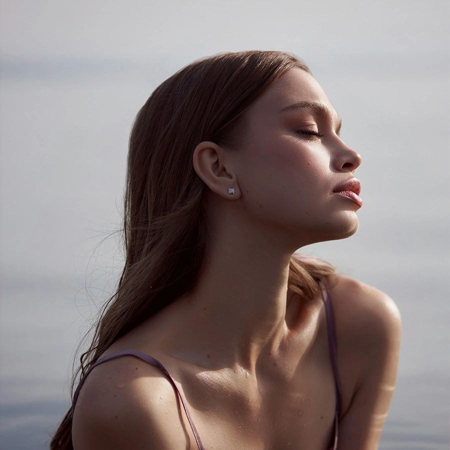 A side profile of a woman with long hair and eyes closed, tilted slightly toward the sun with a calm expression against a misty, light-colored background. - Otoplasty in Beverly Hills, CA