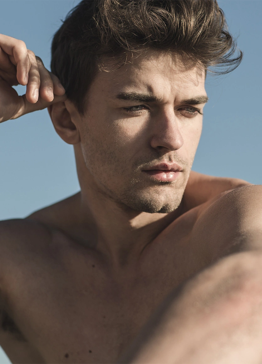 A close-up, low-angle shot of a man with styled brown hair looking downward. He is outdoors under a clear blue sky, with natural sunlight highlighting his facial features and the side of his head. - Otoplasty in Beverly Hills, CA