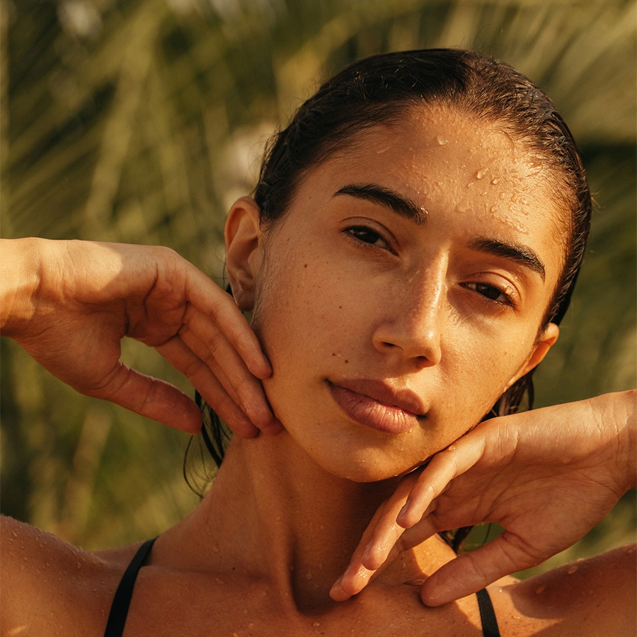 A close-up of a woman with damp, slicked-back hair and water droplets on her face, gently touching her jawline with both hands in golden-hour light. - PRP in Beverly Hills, CA