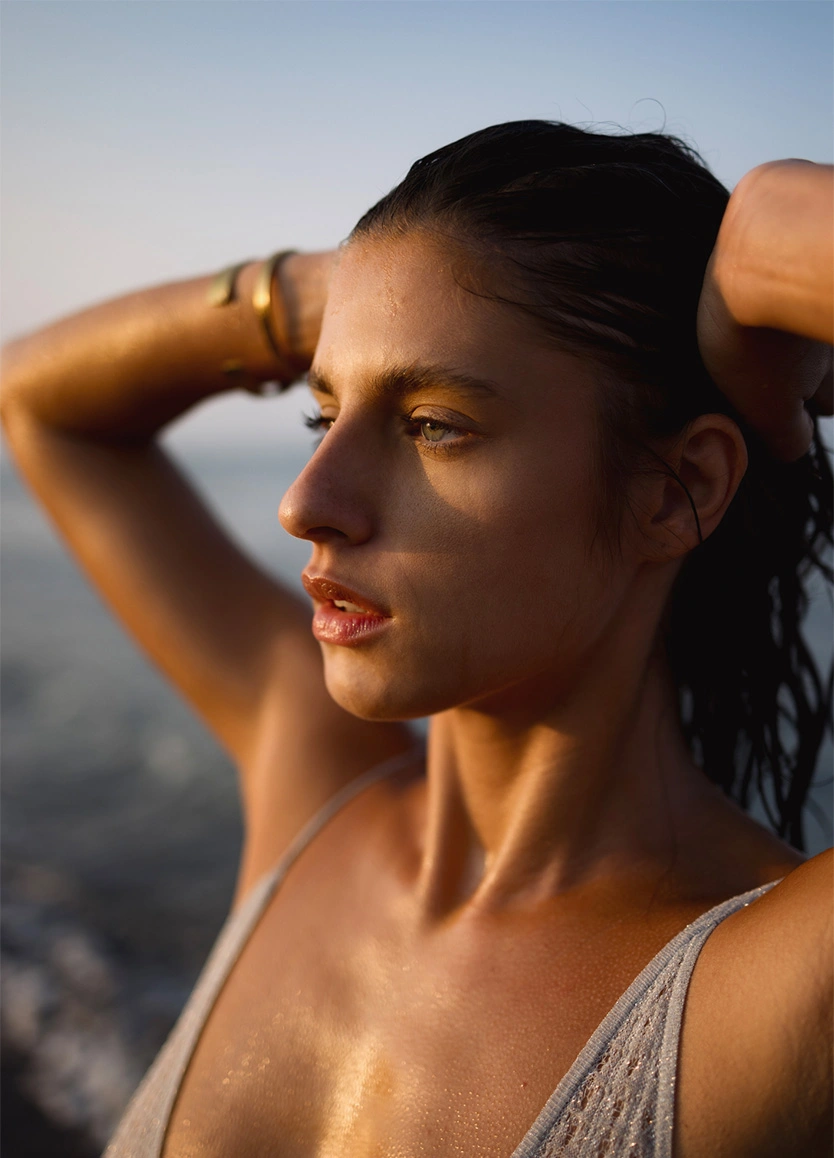 A woman with wet hair and glistening skin looking out toward the ocean during the golden hour. She has her hands raised to her head, and the side-lighting emphasizes her clear complexion and the contour of her face. - PRP injections in Beverly Hills, CA