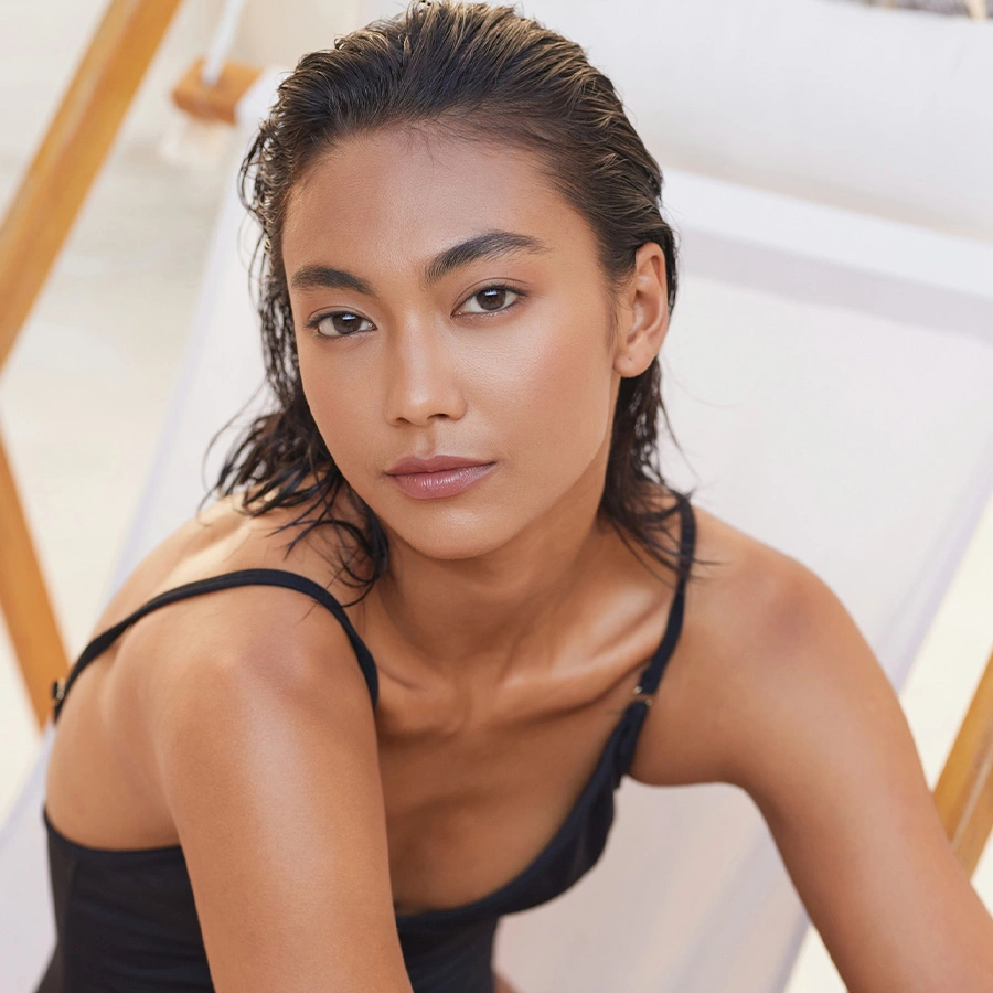 A portrait of a woman with slicked-back dark hair and a black swimsuit, looking directly at the camera while seated in a white lounge chair. - Rhinoplasty in Beverly Hills, CA