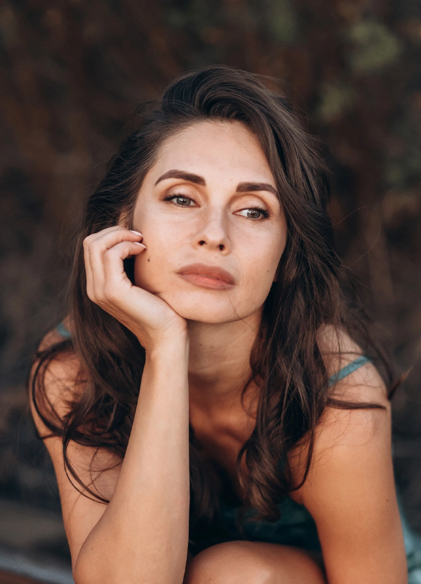 Close-up portrait of a woman with glowing skin resting her chin on her hand. She has long, dark brown hair and is looking off-camera with a thoughtful expression. The background is softly blurred with dark, earthy tones. - SKINVIVE in Beverly Hills, CA