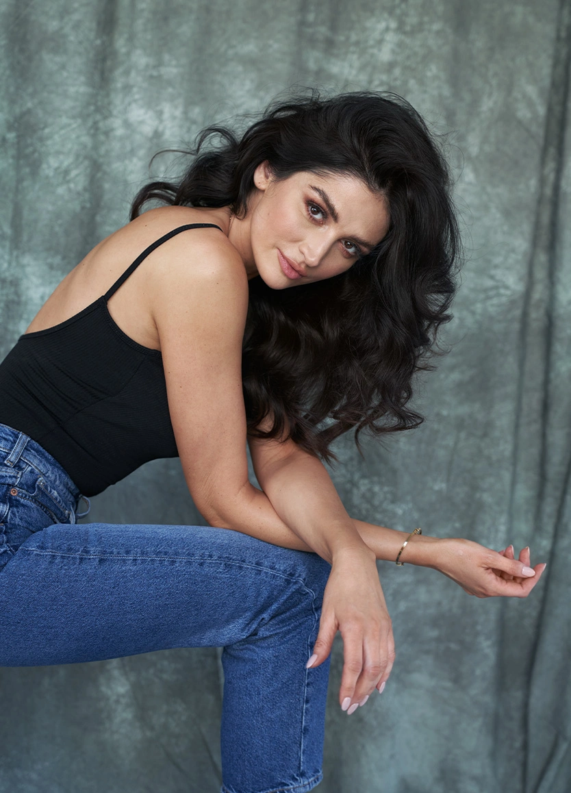 A woman with voluminous, dark wavy hair leaning forward and looking directly into the camera with a confident expression. She is wearing a black spaghetti-strap tank top and blue jeans, posed against a textured gray fabric backdrop. - V1 Peel in Beverly Hills, CA