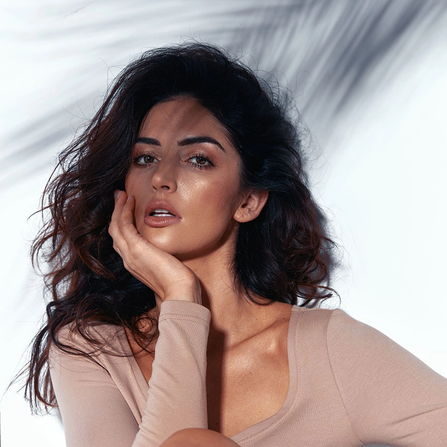 A woman with voluminous, dark wavy hair posing against a white wall. Shadowy patterns from palm leaves are cast across her face and the background. She is wearing a tan long-sleeved top and looking directly at the camera with a poised expression. - Botox in Beverly Hills, CA