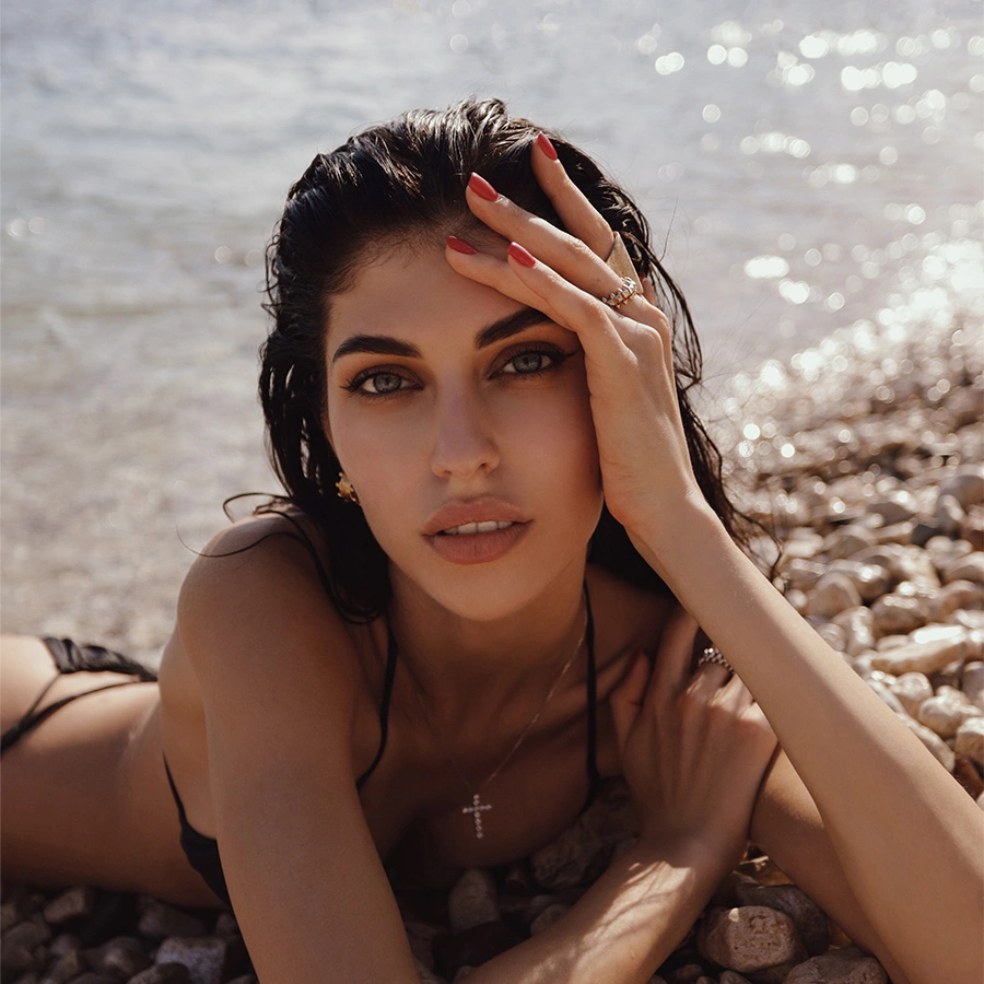 A woman with dark hair and striking blue eyes lying on a pebble beach. She has her hand raised to her forehead, showcasing her well-defined eyebrows and red manicure. The sun-dappled water sparkles in the background. - Brow shaping in Beverly Hills, CA