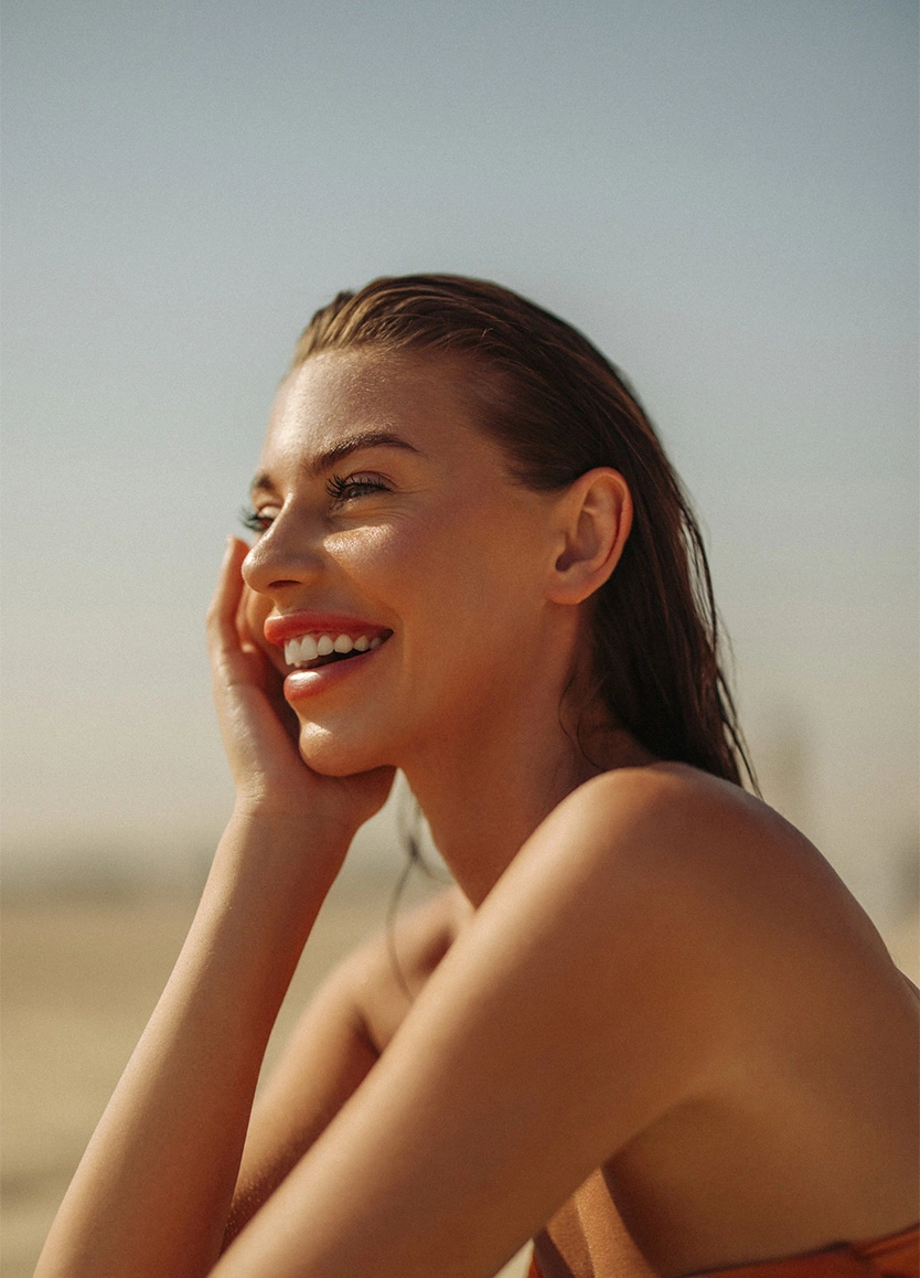 A side-profile view of a woman with slicked-back dark hair laughing joyfully. She is outdoors in bright, natural light, resting her hand against her jaw. The focus is on her perfectly shaped, defined eyebrows and her clear, glowing complexion. - Brow shaping in Beverly Hills, CA