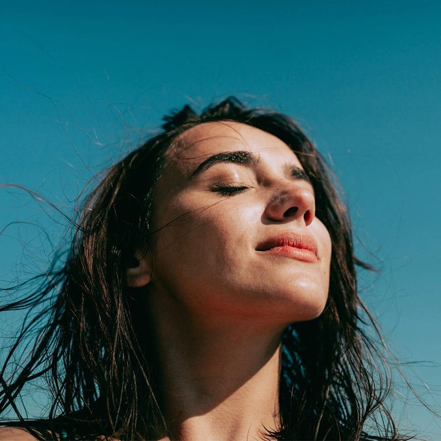 Close-up shot of a woman with her eyes closed and head tilted back, soaking in the sun. Her dark, wind-swept hair is framed against a clear, solid blue sky. The lighting highlights her clear skin and natural features. - Dermaplaning in Beverly Hills, CA