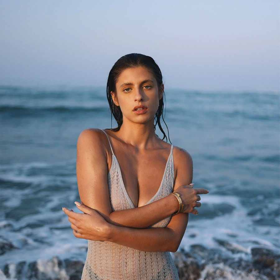 A woman with wet, dark hair pulled back, wearing a grey lace swimsuit. She is standing in front of ocean waves at dusk, with her arms crossed over her chest, looking directly at the camera. - Emsculpt neo in Beverly Hills, CA