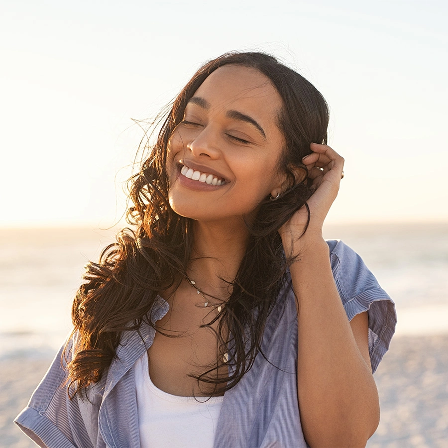 A woman with long, dark curly hair laughing with her eyes closed. She is wearing a white tank top under a light blue button-down shirt. The background is a soft, sandy beach at sunset. - Exilis in Beverly Hills, CA