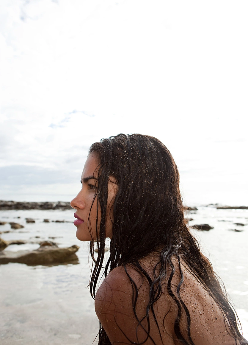 A side profile of a woman with long, damp dark hair standing on a rocky beach under an overcast sky. Her skin and hair are lightly dusted with sand, and she looks out toward the ocean. The natural light emphasizes the smooth texture of her profile and the firm contours of her neck and jaw. - Exilis in Beverly Hills, CA