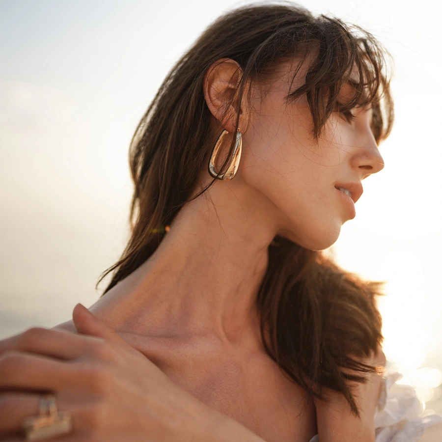 A soft-focus side profile of a woman with dark hair tucked behind her ear, revealing a large, thick gold hoop earring. She is touching her shoulder with her hand, and the background is a bright, hazy outdoor light. - Facial treatment in Beverly Hills, CA