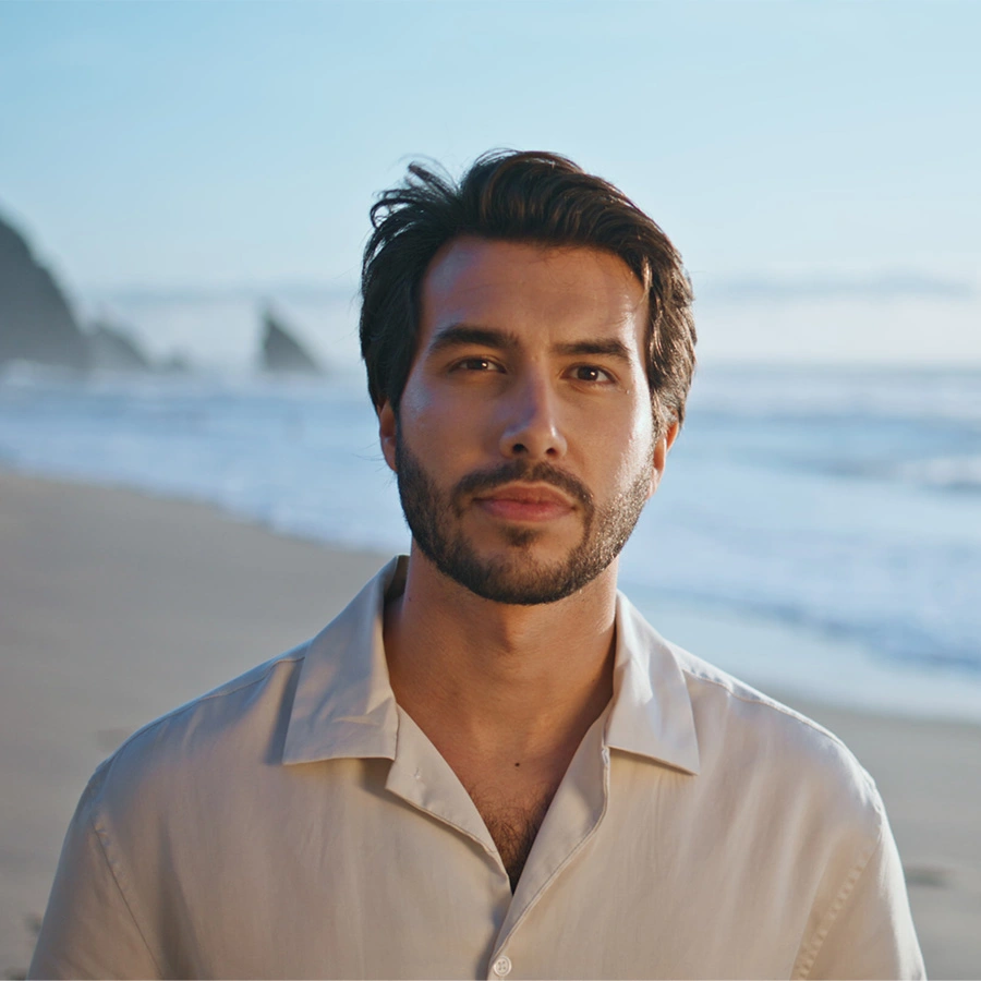 A front-facing portrait of a man with dark hair, a well-groomed beard, and a tan complexion. He is wearing a light-colored, button-down linen shirt. The background is a soft-focus beach with waves and distant rock formations. - Hair restoration in Beverly Hills, CA