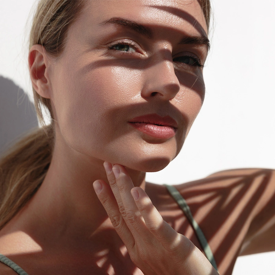 A close-up of a woman's face with smooth, glowing skin. She has her hand gently touching her chin. Shadows from palm fronds are cast across her face and a white wall in the background, creating a high-contrast, sunny aesthetic. - Hydrafacial in Beverly Hills, CA