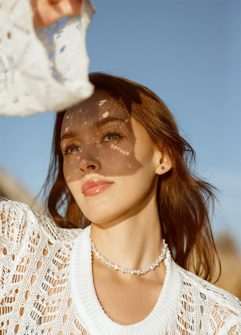 A close-up, low-angle portrait of a woman with long auburn hair looking upward with a serene expression. She is wearing a white crochet top and a delicate pearl necklace. A lacy fabric is held above her, casting intricate, dappled shadows across her forehead and eyes. The lighting is warm and golden against a clear blue sky, highlighting her defined jawline and glowing skin. - Jawline contouring in Beverly Hills, CA