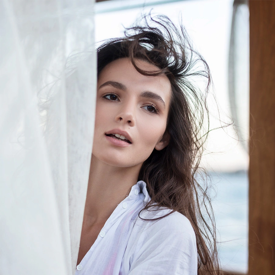 A woman with long dark hair peeks from behind a sheer white curtain, looking off into the distance with a neutral expression. - Kybella in Beverly Hills, CA