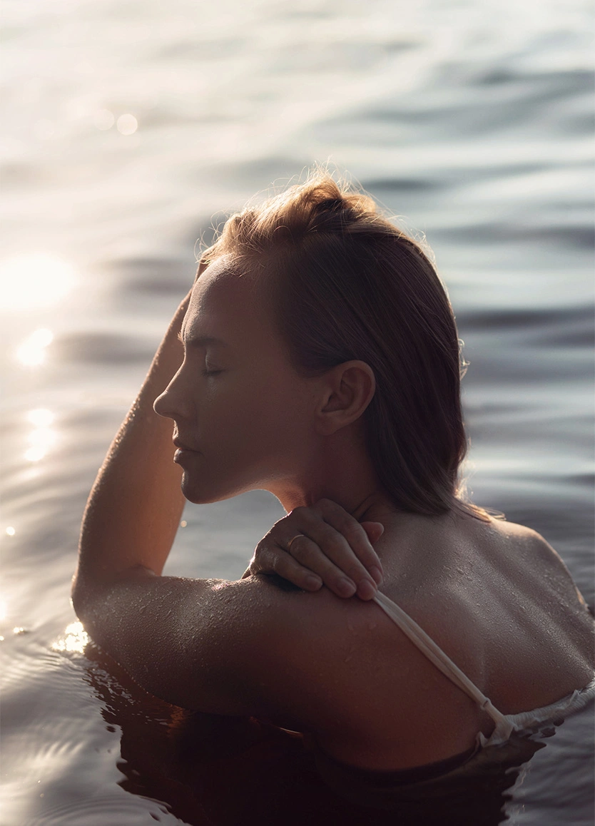 A woman submerged in calm water up to her shoulders, her profile illuminated by a bright, shimmering sun reflection on the water’s surface. Her eyes are closed, and her skin appears wet and radiant. - microneedling in Beverly Hills, CA